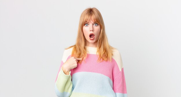 pretty red head woman looking shocked and surprised with mouth wide open, pointing to self