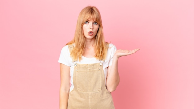 Pretty red head woman looking surprised and shocked, with jaw dropped holding an object