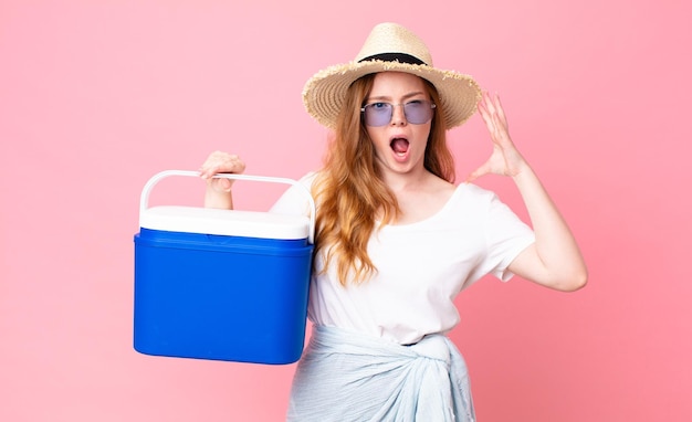 Bella donna con la testa rossa che urla con le mani in aria e tiene in mano un frigorifero portatile da picnic