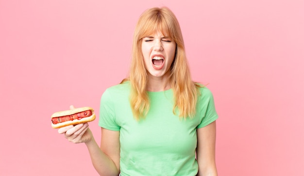 Pretty red head woman shouting aggressively, looking very angry and holding a hot dog