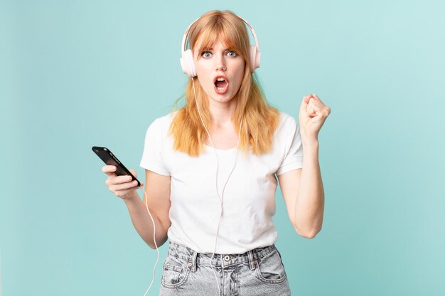 Pretty red head woman shouting aggressively with an angry expression and listening music with headphones