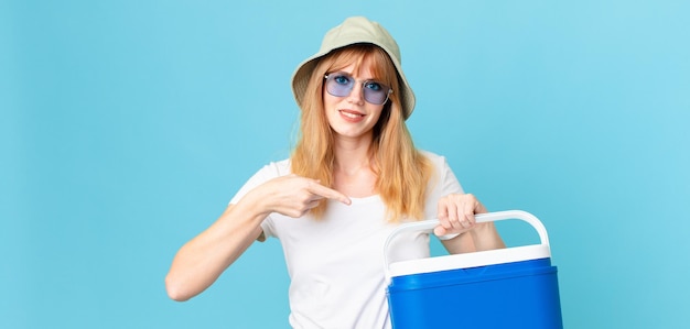 Pretty red head woman smiling cheerfully, feeling happy and pointing to the side and holding a portable refrigerator. summer concept