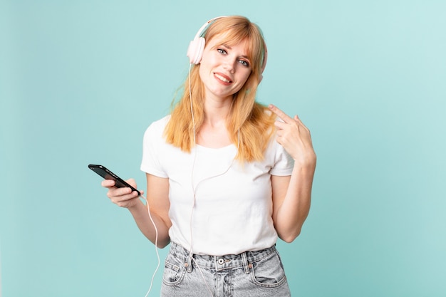 Pretty red head woman smiling confidently pointing to own broad smile and listening music with headphones