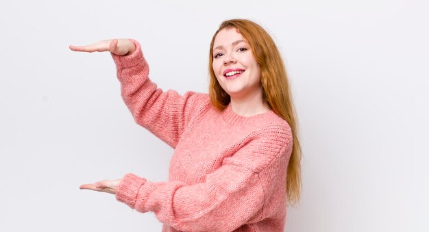 Pretty red head woman smiling feeling happy positive and satisfied holding or showing object or concept on copy space