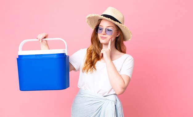 Pretty red head woman smiling happily and daydreaming or doubting and holding a picnic portable refrigerator