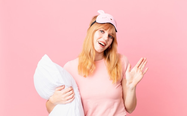 Pretty red head woman smiling happily, waving hand, welcoming and greeting you. wearing pajamas and holding a pillow