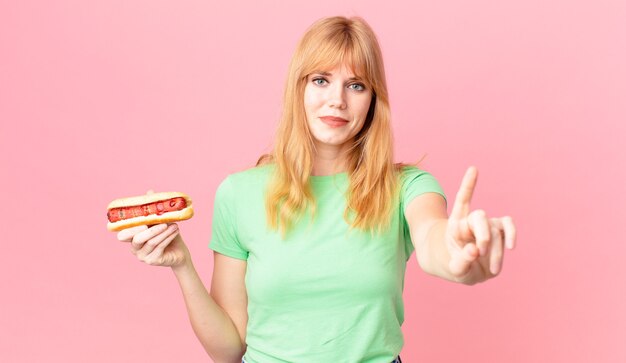 Pretty red head woman smiling and looking friendly, showing number one and holding a hot dog