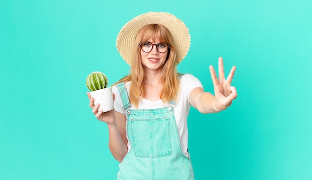 Bella donna dalla testa rossa che sorride e sembra amichevole, mostra il numero tre e tiene in mano un cactus in vaso. concetto di contadino