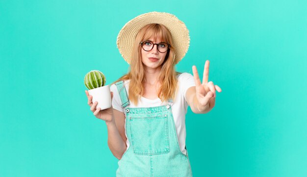 Pretty red head woman smiling and looking friendly, showing number two and holding a potted cactus. farmer concept