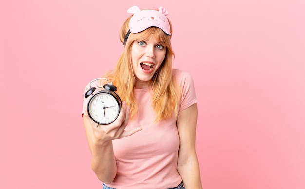 Pretty red head woman wearing pajamas and holding an alarm clock wake up concept