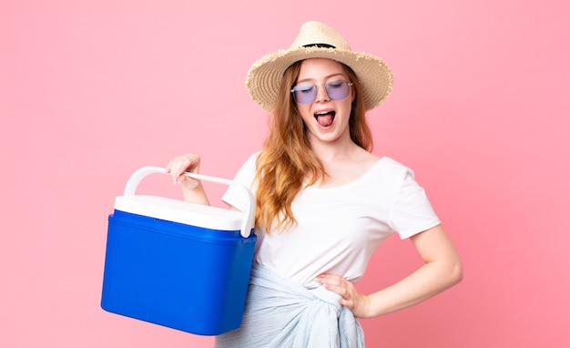 Pretty red head woman with cheerful and rebellious attitude, joking and sticking tongue out and holding a picnic portable refrigerator