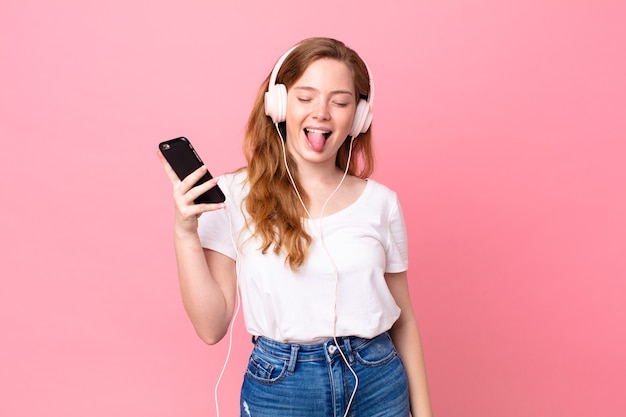pretty red head woman with cheerful and rebellious attitude, joking and sticking tongue out with headphones and smartphone