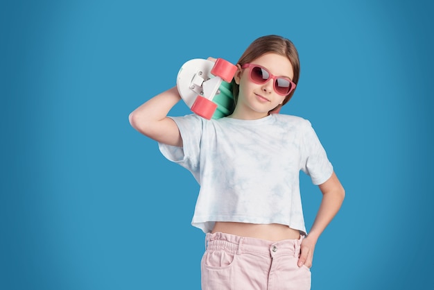 Pretty relaxed teenager in sunglasses, white t-shirt and jeans holding skateboard on shoulder while standing in front of camera