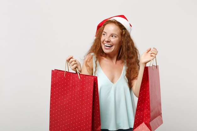 Pretty roodharige santa meisje in kerstmuts geïsoleerd op een witte achtergrond. gelukkig nieuwjaar 2020 viering vakantie concept. bespotten kopie ruimte. houd de pakkettas met geschenken of aankopen vast na het winkelen.