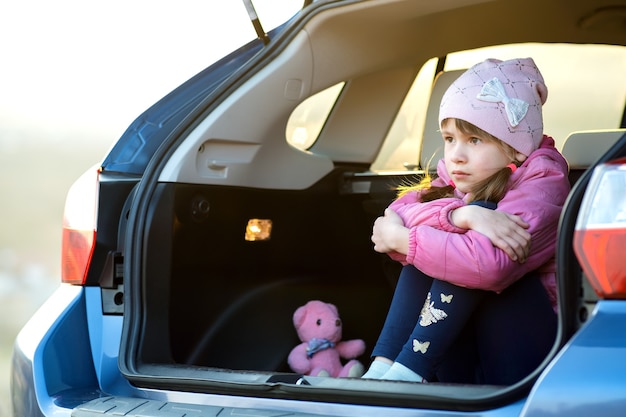 Pretty sad child girl sitting alone in a car trunk with a pink toy teddy bear.