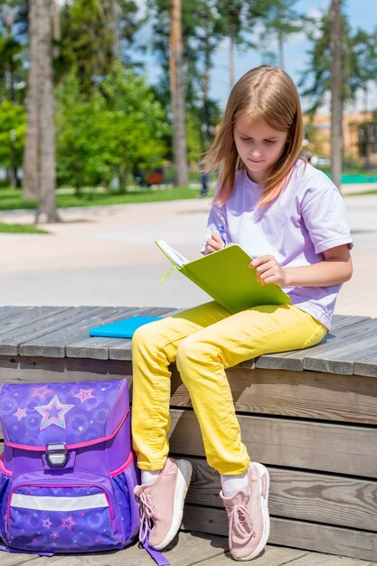 Pretty school girl sitting on the bench in public park and making notes to her note book