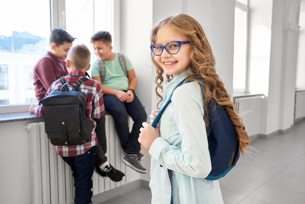 Pretty school girl with blonde long hair and backpack on shoulder standing on corridor near classroom.