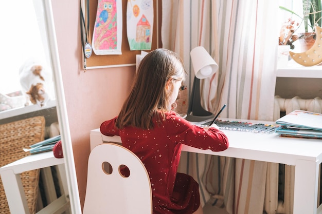 Pretty school girl with long hair in red dress draws with pencil at table in childrens room at home