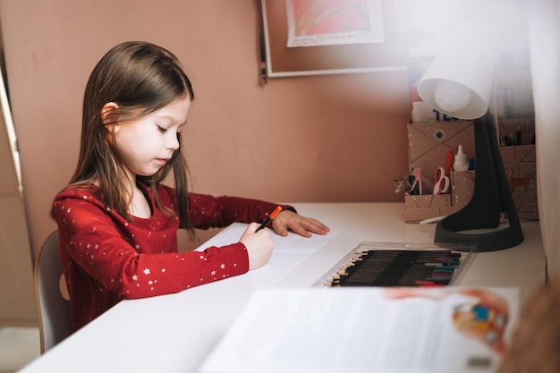 Pretty school girl with long hair in red dress draws with pencil at table in childrens room at home