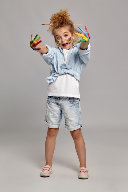 Pretty schoolgirl having a brush in her chic curly blond hair, wearing in a blue shirt and white t-shirt. She is showing her painted hands and open her mouth, on a gray background.