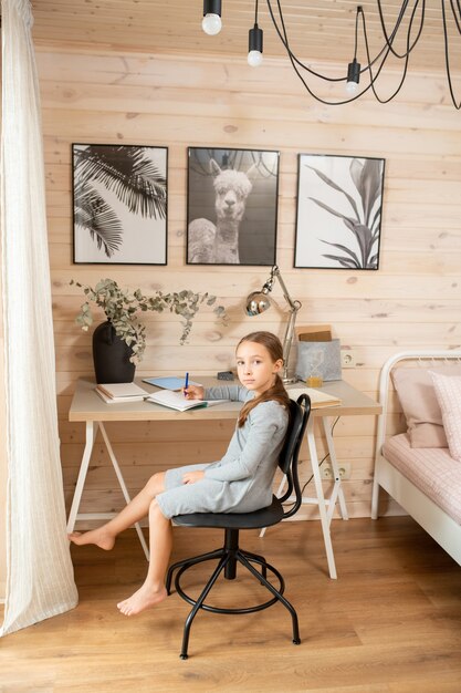 Pretty schoolgirl in homewear sitting on chair by table against wooden wall with pictures and looking at you while doing homework in bedroom