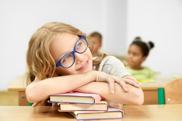 Pretty schoolgirl sitting at desk in classroom, leaning head on stack of books. 