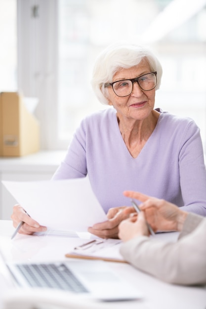 Pretty senior woman in eyeglasses and casualwear holding insurance document while discussing its terms and conditions with agent