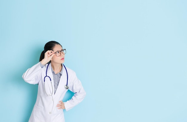 pretty seriously female hospital doctor holding glasses looking at medical empty area thinking healthy problem isolated on blue wall background.