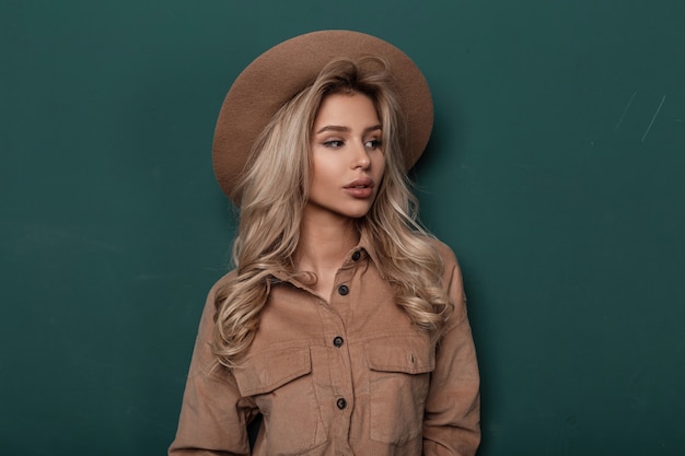 Pretty sexy young woman in an elegant beige hat in a stylish shirt with blond curly blond hair stands in the studio near a green vintage wall