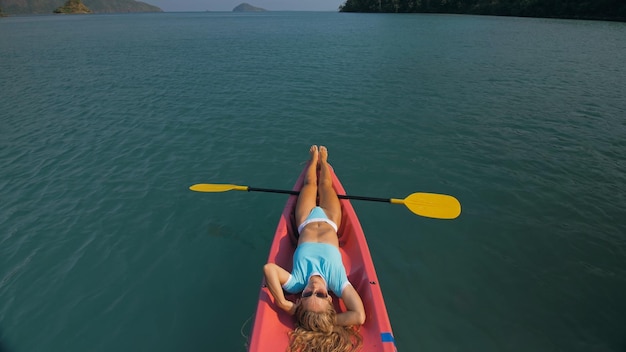 Pretty slim woman in elegant swimsuit lies in pink plastic kayak drifting on azure ocean