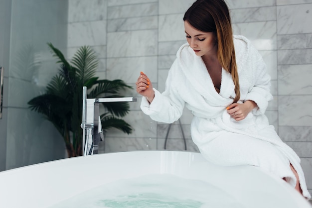 Pretty slim woman wearing bathrobe sitting on edge of bathtub filling up with water.