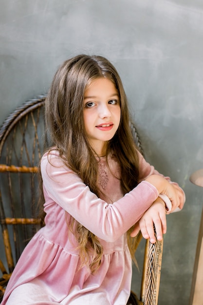 Pretty small girl in beautiful pink dress sits on the wooden chair and looks dreamy, beauty and fashion concept, indoor portrait