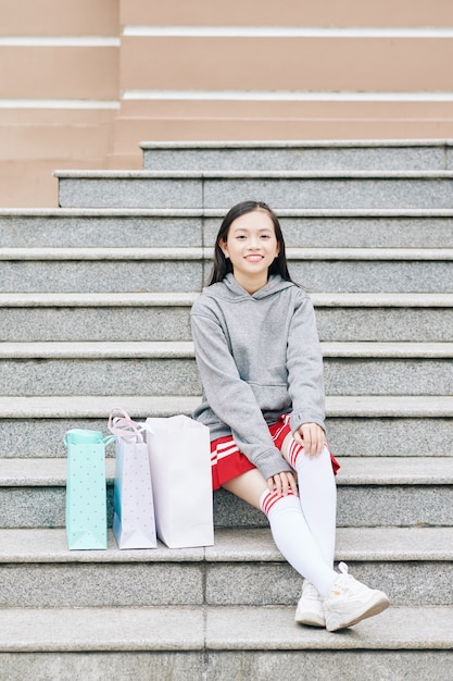 Довольно улыбается азиатская девочка-подросток, сидящая на ступеньках рядом с сумками для покупок