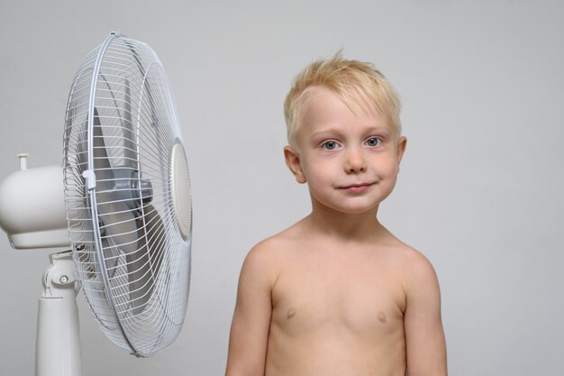 Il ragazzo biondo abbastanza sorridente con il torso nudo sta vicino ad un ventilatore. concetto di estate