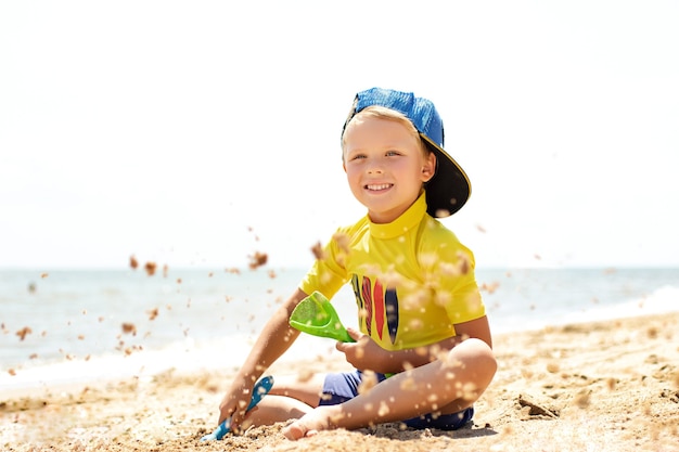 Довольно улыбающийся мальчик сидит на пляже и играет с песком. Концепция летнего отдыха.