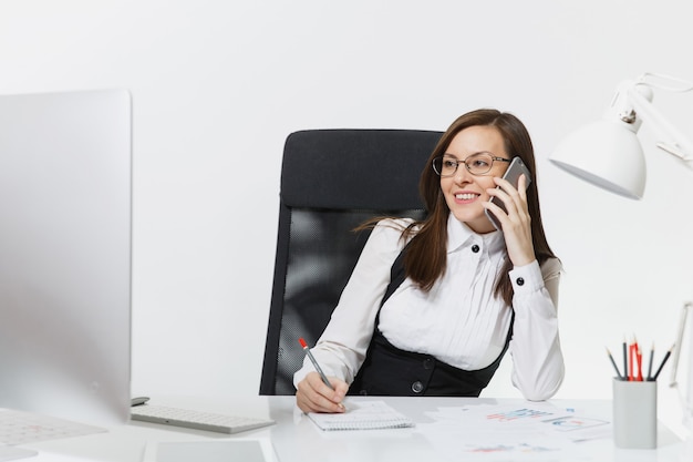 Довольно улыбающаяся деловая женщина в костюме сидит за столом, работает за современным компьютером с документом в легком офисе, разговаривает по мобильному телефону, ведет приятную беседу