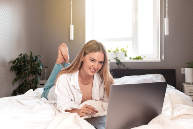 La ragazza caucasica abbastanza sorridente giace sul letto in camera da letto e utilizza il laptop per il lavoro a distanza. comunicazione, tecnologia, concetti di lavoro a distanza in appartamento interno grigio