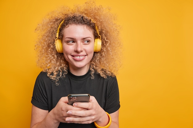 Pretty smiling curly haired young woman looks away happily listens music uses smartphone enjoys good sound quality wears casual black t shirt isolated over yellow wall with blank space