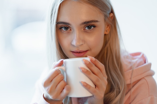 A pretty smiling girl enjoying a cup of coffee or milk while sitting by the blurred window glass. Wearing a casual pink, pale parka. Close up white mug in woman hands near lips.