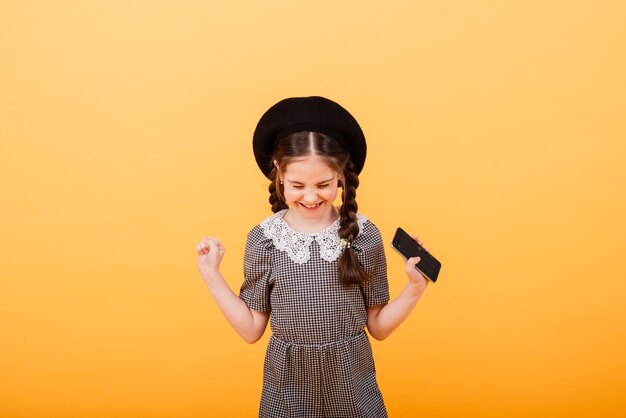 Pretty smiling girl is holding smartphone. Cute child on yellow background, modern children concept.