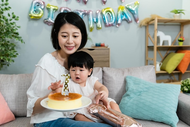 pretty smiling japanese mother is holding and looking at the cake in hands while her cute baby girl reaching for the candle with curiosity at a birthday party.