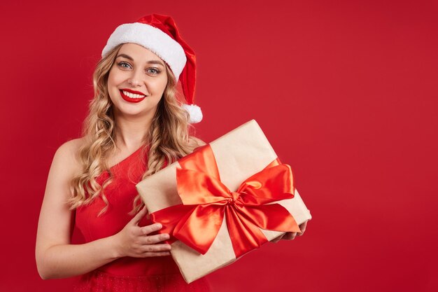 Pretty smiling lady in a red dress wearing a santa hat holding a gift box with a bow