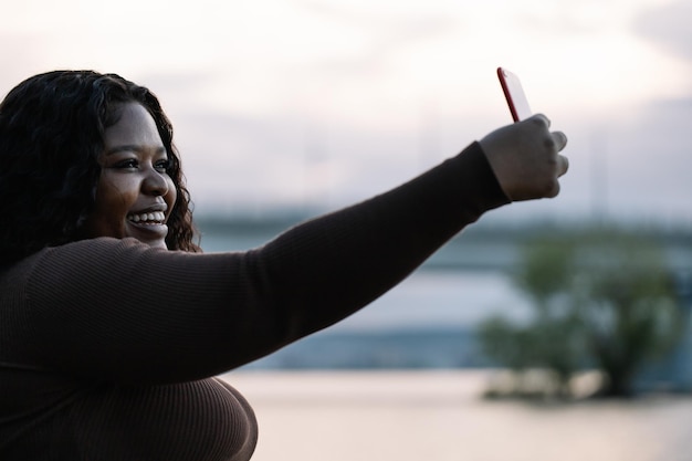 Pretty smiling plump interracial woman in dress take selfie video call photo smartphone pose against sky Acceptance