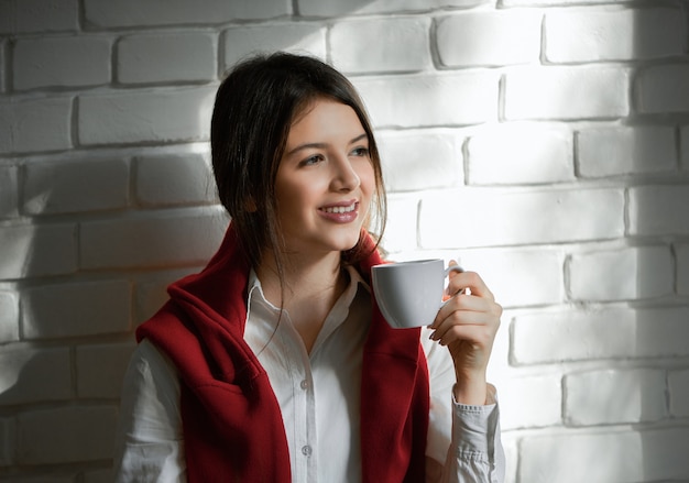 Studente abbastanza sorridente che beve il caffè al mattino. avere capelli neri corti e trucco leggero. indossare casual camicia bianca e cardigan rosso. sentirsi bene, felice.