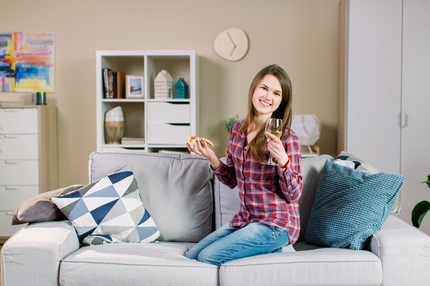 Pretty smiling woman in jeans and casual shirt, drinking wine and eating pizza,while sitting on gray sofa at home