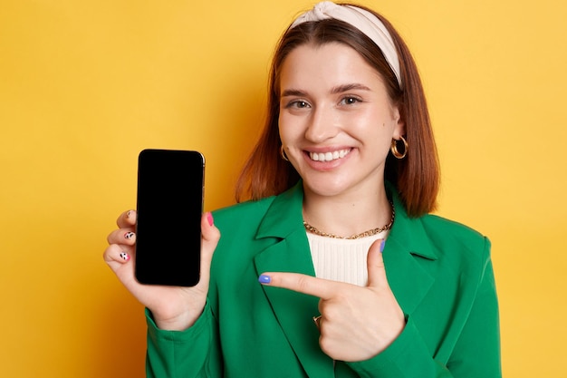 Pretty smiling woman wearing green jacket posing isolated over yellow background pointing at smartphone with empty display empty space for advertisement