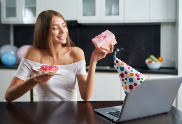 Pretty smiling woman with donut and gift box in hands having video call on modern laptop