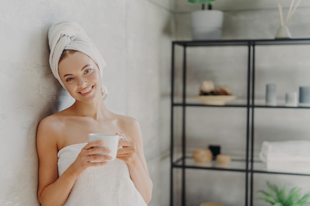 Pretty smiling woman with healthy skin wrapped in white towel stands glad indoor drinks coffee has positive face expression poses indoor People spa pampering skin care and wellness concept