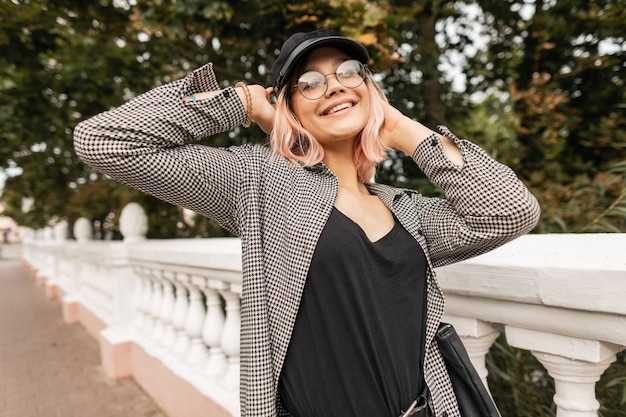 Довольно улыбающаяся молодая девушка с красивым эмоциональным лицом в модных очках в стильной одежде, наслаждаясь прогулкой в парке