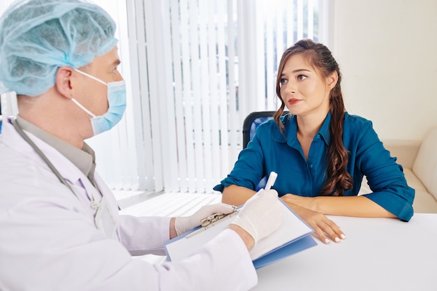 Pretty smiling young woman listening to recommendations of doctor in medical mask and rubber gloves